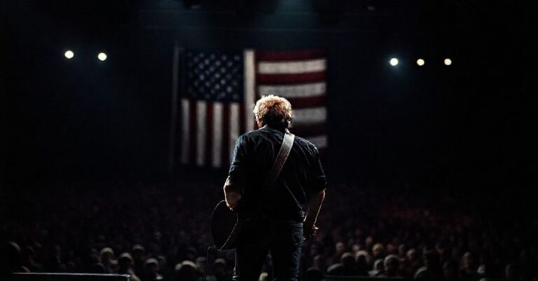 Bruce Springsteen na scenie w cieniu spotów, z gitarą w dłoni, przeciwko tłu amerykańskiej flagi