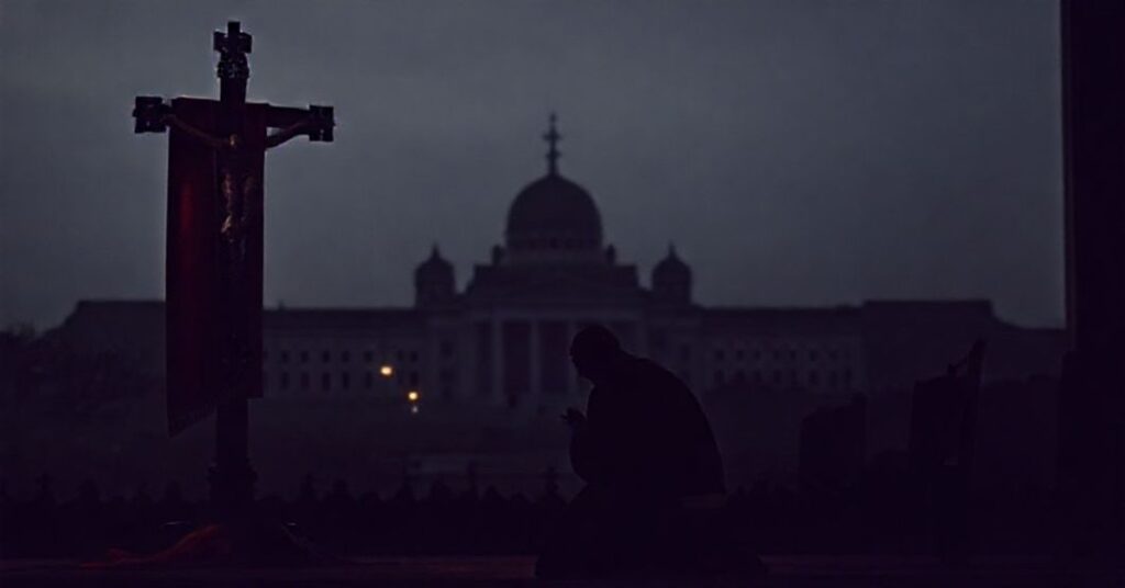 Prawosławny kapłan modli się przed krzyżem z flagą Gruzji w tle; symbolizuje duchowy kryzys kraju i nadzieję na Boże działanie.