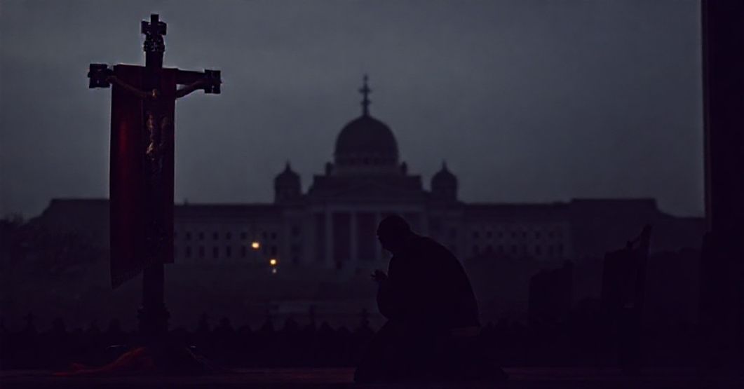 Prawosławny kapłan modli się przed krzyżem z flagą Gruzji w tle; symbolizuje duchowy kryzys kraju i nadzieję na Boże działanie.