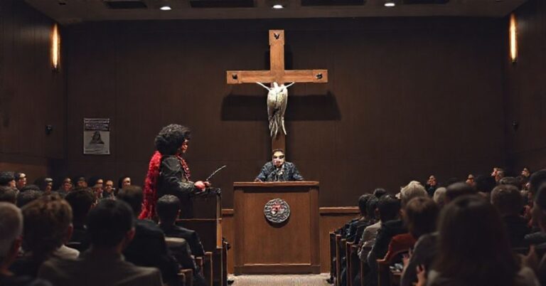 Zdjęcie przedstawiaje scenę w auditorium Uniwersytetu Harvarda, gdzie profesor drag queen Kareem Khubchandani (pseudonim "LaWhore Vagistan") wygłasza wykład w prowokującym stroju kobiecym. Tło ilustruje upadek moralny uczelni.