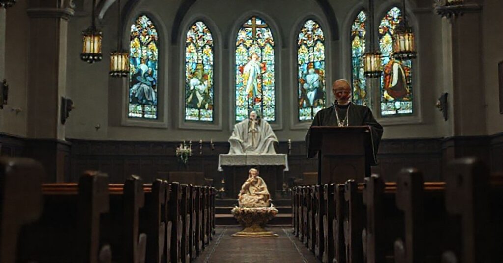 Wnętrze kościoła z amboną, gdzie fałszywy biskup Krzysztof Chudzio wygłasza herezyjne kazanie. Na pierwszym planie wizerunek Dzieciątka Jezus otoczonego ofiarami Votyw. Scena jest przygaszona i niepokojąca.
