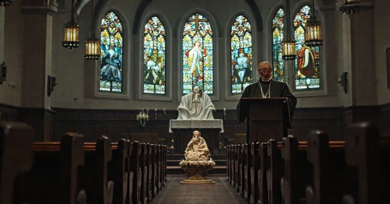 Wnętrze kościoła z amboną, gdzie fałszywy biskup Krzysztof Chudzio wygłasza herezyjne kazanie. Na pierwszym planie wizerunek Dzieciątka Jezus otoczonego ofiarami Votyw. Scena jest przygaszona i niepokojąca.