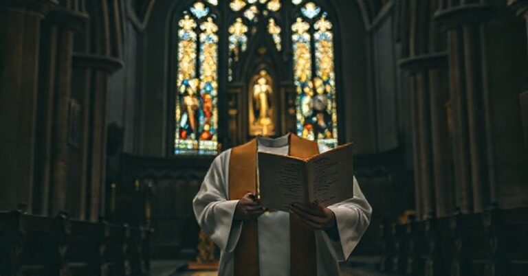 Sobótni kapłan w tradycyjnych szatach stoi w ciemnym kościele przed ołtarzem z książką Alberta Cohena.