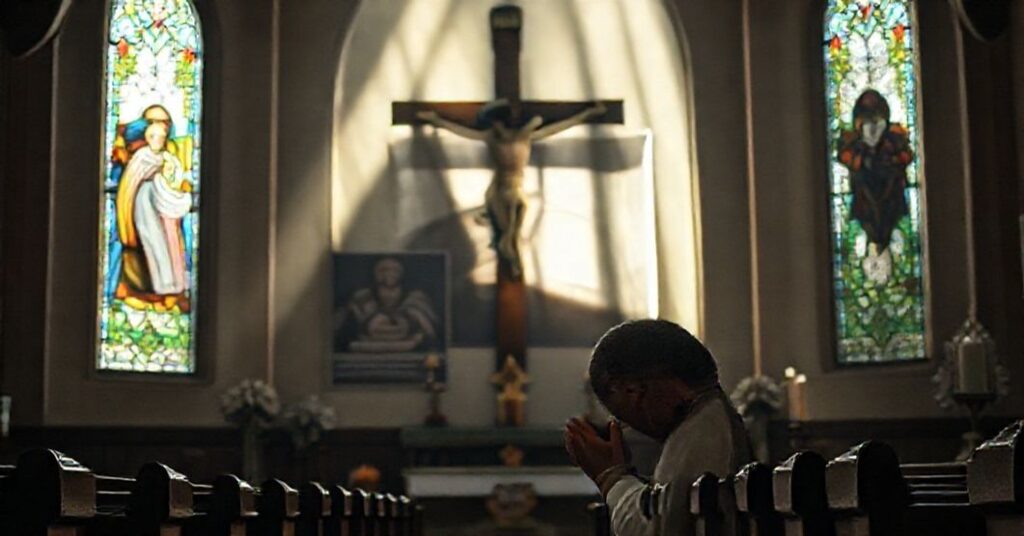 Ksiądz katolicki modli się przed krzyżem w kościele z witrażem przedstawiającym Zwiastowanie. Tło symbolizuje secolaryzację ruchu "miast-sanktuariuszów".