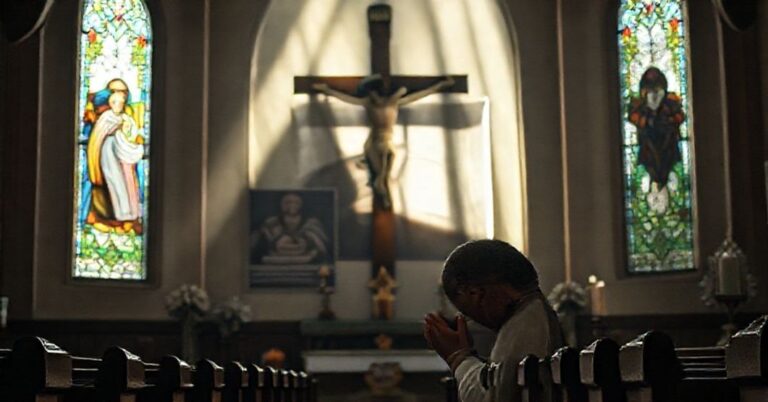 Ksiądz katolicki modli się przed krzyżem w kościele z witrażem przedstawiającym Zwiastowanie. Tło symbolizuje secolaryzację ruchu "miast-sanktuariuszów".