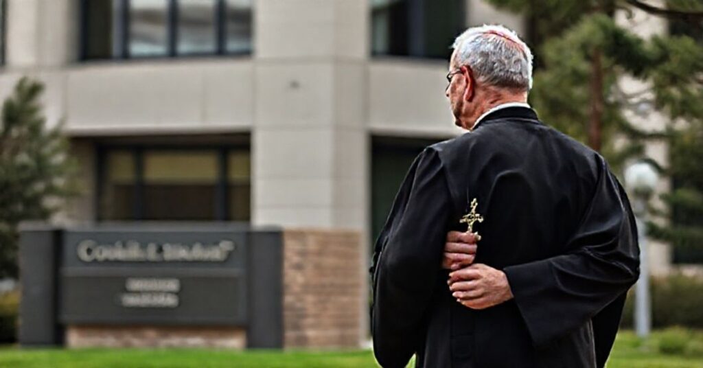 Katolicki lekarz w tradycyjnym ubraniu duchownego stoi przed budynkiem Uniwersytetu Kolorado, trzymając krucyfiks. Scena ukazuje konflikt wiary z władzą państwową.