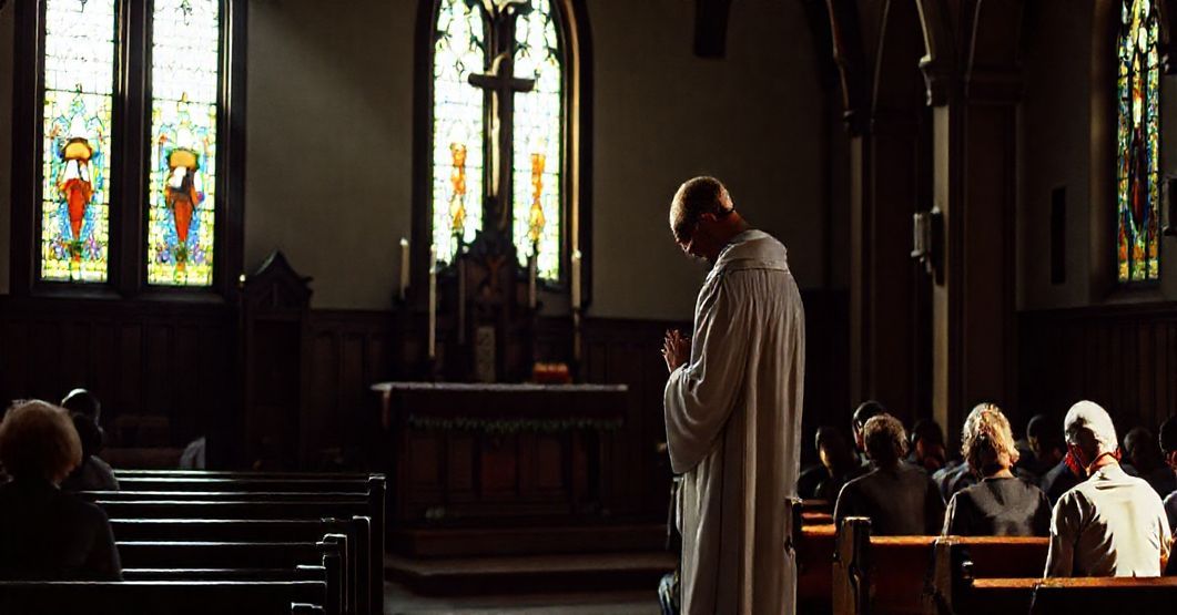 Ciemna wnętrza kościoła z tradycyjnym ołtarzem i modlącym się księdzem w stroju liturgicznym. Skupieni parafianie w refleksji.