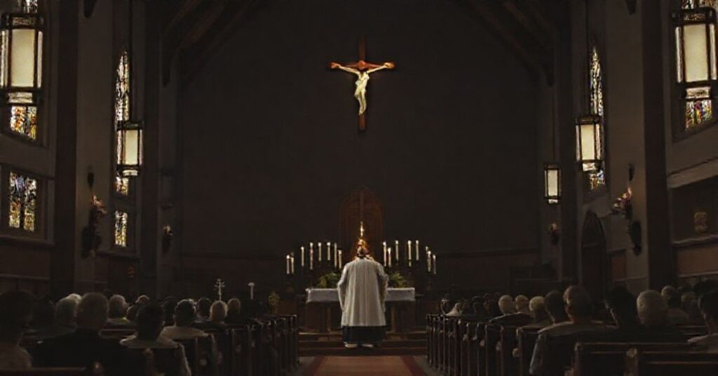 Wnętrze tradycyjnego kościoła katolickiego z krzyżem jako centralnym elementem i kapłanem w sutannie stojącym przed ołtarzem.