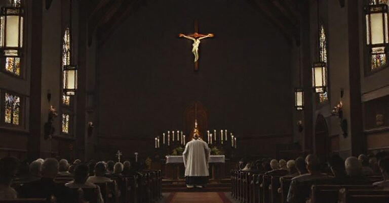 Wnętrze tradycyjnego kościoła katolickiego z krzyżem jako centralnym elementem i kapłanem w sutannie stojącym przed ołtarzem.