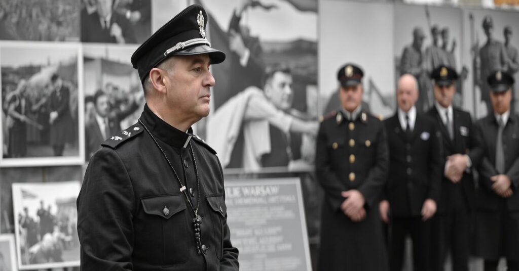 Tradycyjny ksiądz w mundurze wojskowym przed pomnikiem powstańczym, otoczony zdjęciami z Powstania Warszawskiego. Na twarzy modlitwa i kontemplacja. W tle widoczni świeccy urzędnicy.