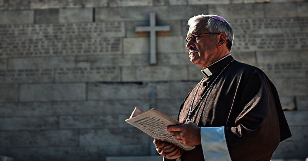 Ksiądz w tradycyjnych szatach kościelnych stoi przed monumentem Instytutu Jad Waszem, trzymając encyklikę Piusa XI. Tło przedstawia ciemne mury z hebrajskimi napisami.