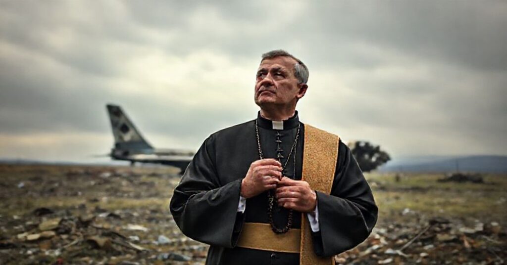 Ksiądz w tradycyjnych szatach modli się przed zniszczonym krajobrazem Krymu z widocznym zniszczonym rosyjskim samolotem w tle.