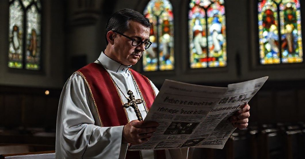 Ksiądz w tradycyjnym stroju liturgicznym trzyma krzyż podczas rezonansowej modlitwy w świetle witraży kościelnych.