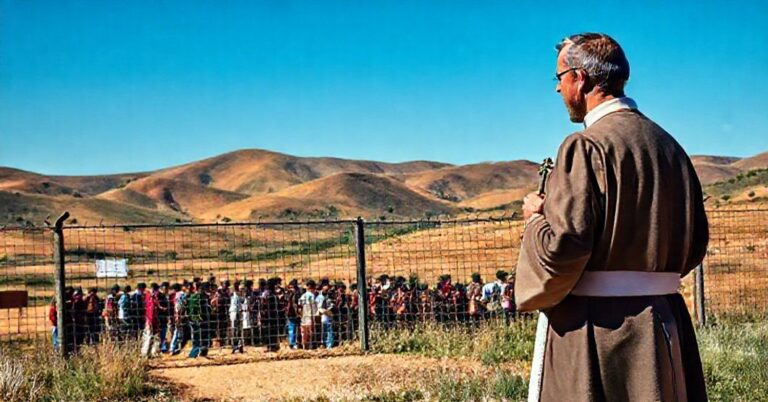 Ksiądz w tradycyjnych szatach stoi przed zamkniętą granicą narodową, trzymając krzyż i patrząc poważnie na grupę nielegalnych imigrantów zgromadzonych po drugiej stronie ogrodzenia.