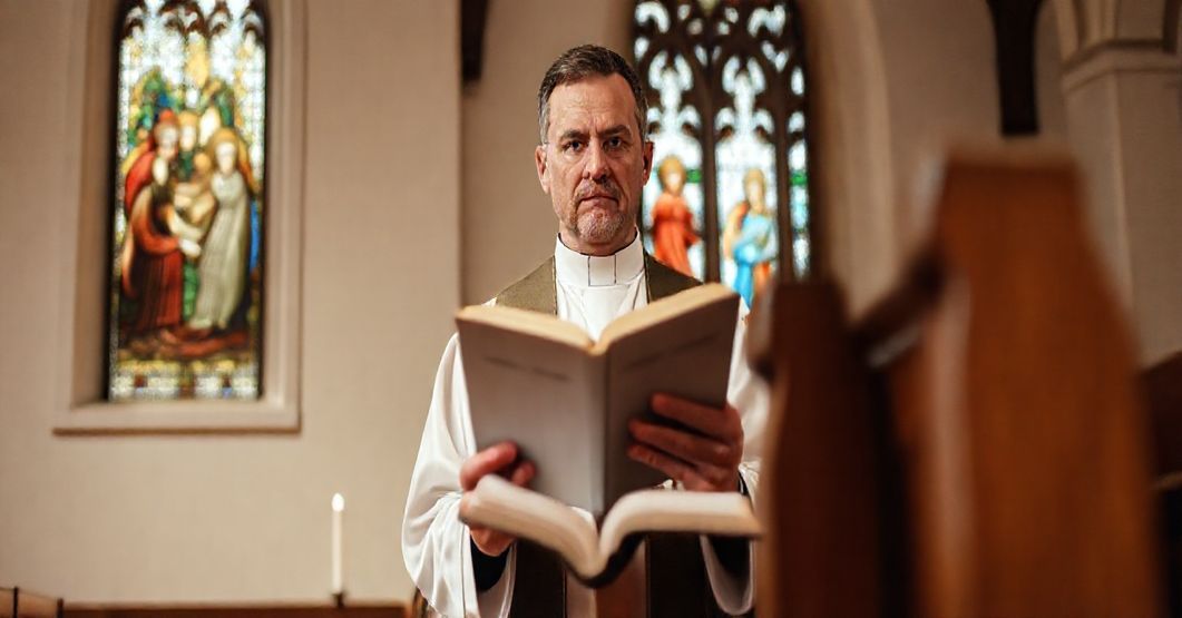 Ksiądz w tradycyjnym stroju liturgicznym trzyma Biblię w kościele, z witrażem przedstawiającym Świętą Rodzina na tle