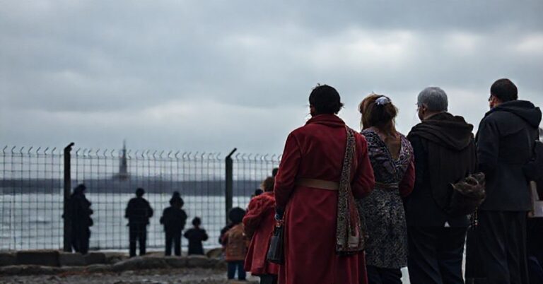 Grupa uchodźców stoi przy kanale La Manche, patrząc na zamkniętą granicę Wielkiej Brytanii. Scena jest poważna i smutna, z akcentem na problem migracji z katolickiego punktu widzenia.