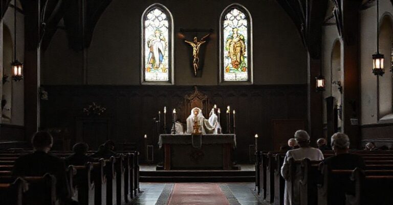 Wnętrze kościoła w Szklarce Radnickiej podczas mszy. Stary ołtarz z krucyfiksem i zapalonymi świecami. Kleryk w tradycyjnej szacie liturgicznej. Wierni modlący się w ciszy.