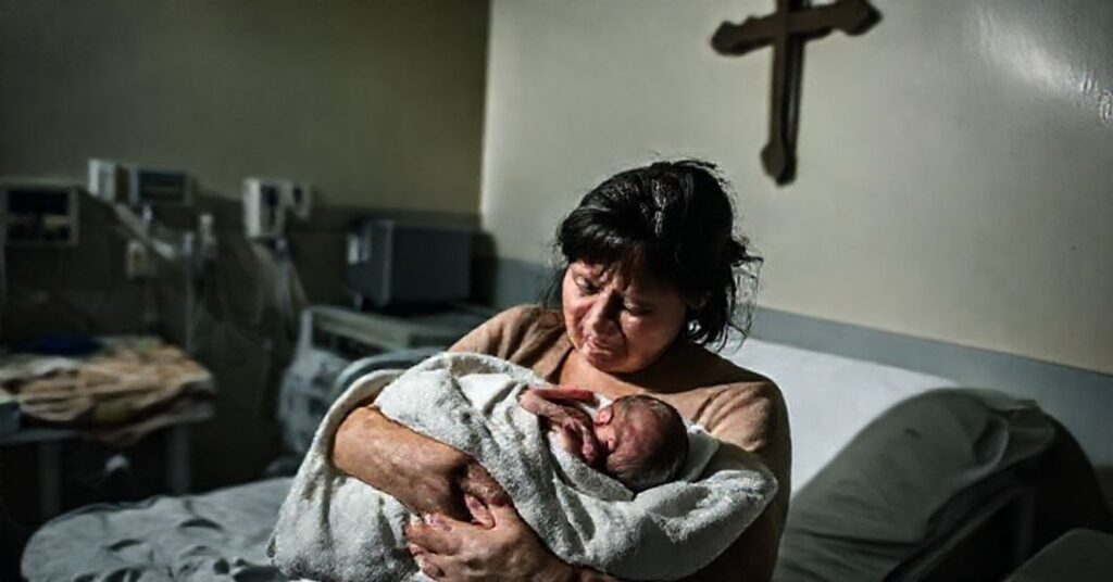 Scena szpitalna w Nowej Zelandii z matką trzymającą martwe dziecko po aborcji, w tle sprzęt medyczny i krzyż.