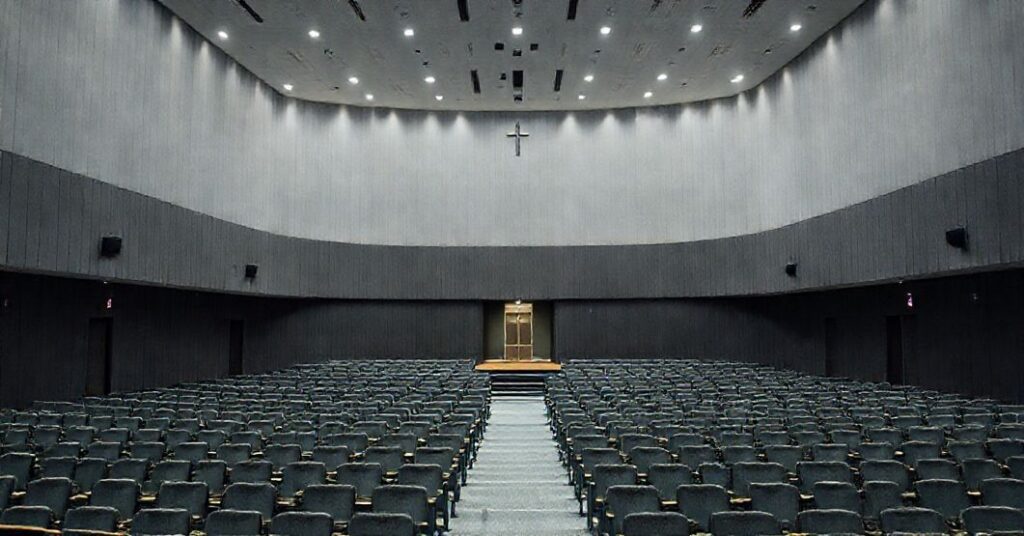 Obraz przedstawiający nowoczesne seminarium w Tarnowie z modernistyczną architekturą. W centrum uwagi znajduje się duża sala konferencyjna przeznaczona do wydarzeń świeckich, a w tle mała, prawie ukryta kaplica. Scena jest pozbawiona katolickich symboli, podkreślając brak formacji doktrynalnej. Atmosfera wydaje się być bardziej biurokratyczna niż duchowa.