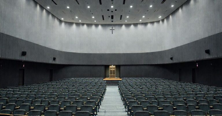 Obraz przedstawiający nowoczesne seminarium w Tarnowie z modernistyczną architekturą. W centrum uwagi znajduje się duża sala konferencyjna przeznaczona do wydarzeń świeckich, a w tle mała, prawie ukryta kaplica. Scena jest pozbawiona katolickich symboli, podkreślając brak formacji doktrynalnej. Atmosfera wydaje się być bardziej biurokratyczna niż duchowa.