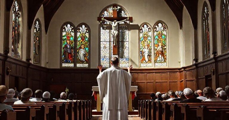 Tradycyjny wnętrze kościoła katolickiego z ks. w białej szacie modlącego się przed ołtarzem, tłem duży krzyż i wierni na kolanach.