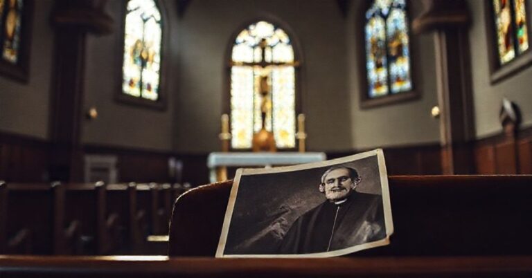 Wnętrze tradycyjnego kościoła katolickiego z ołtarzem i krzyżem Chrystusowym na tle witraży. Zdalnie widoczny zdjęcie ks. Jerzego Popiełuszki na ławce.