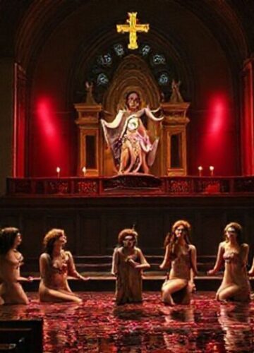 Wnętrze katolickiego kościoła z rzeźbą Matki Boskiej Bolesnej w pierwszym planie, patrzącą z żalem na scenę teatralną z aktorkami w nieprzyzwoitych kostiumach i sztucznym kałem.