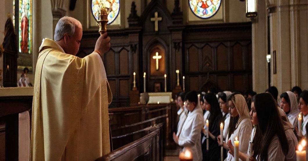 Tradycyjna msza katolicka z podniesionym Hostią podczas konsekracji w klasycznym kościele z modlącymi się młodymi ludźmi.